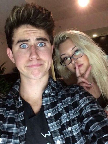 nash wearing a check shirt with stassie in the back wearing glasses with her finger on her lips 